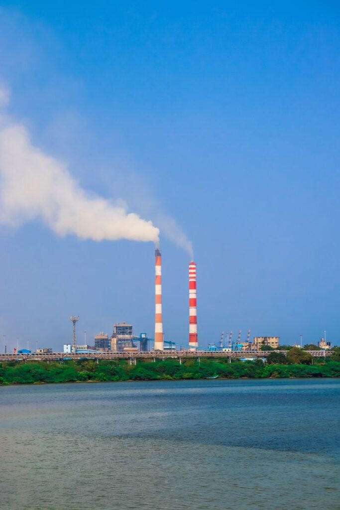 pexels-photo-20220788-20220788 Industrial scene with chimneys emitting smoke by a lake under a clear blue sky.